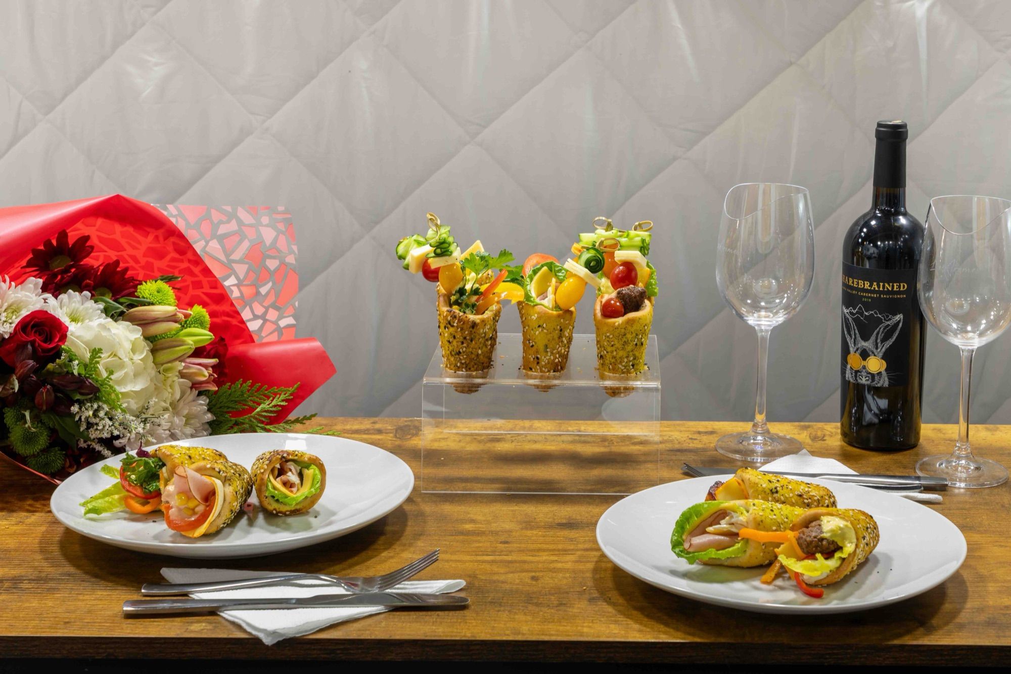 A table set for two with filled BreadKones on plates, mini cones on an acrylic stand, flowers, and red wine