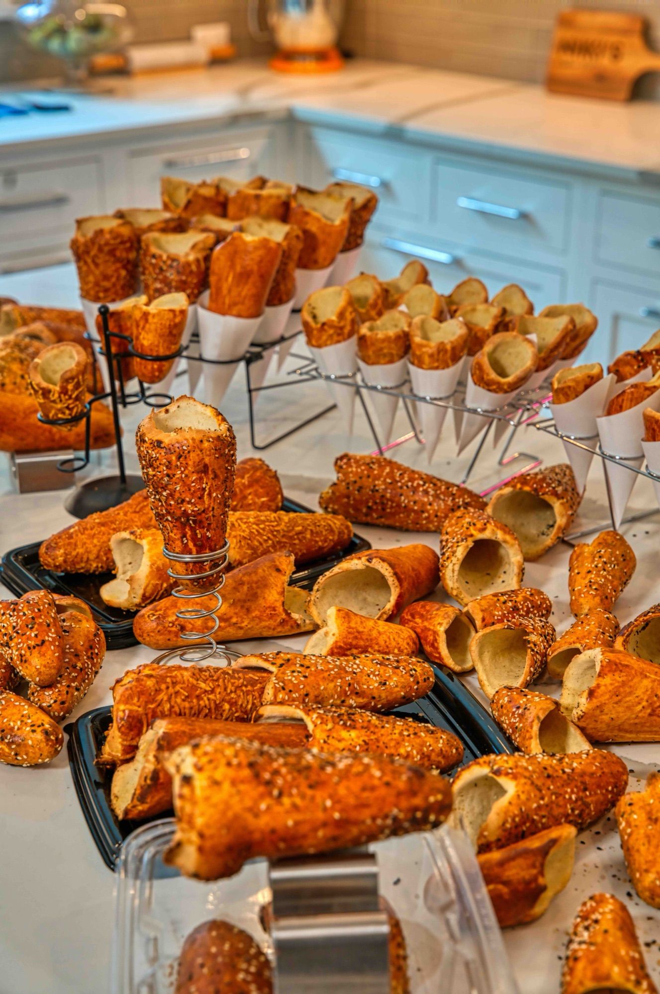 A catering spread of BreadKones in paper cones on wire racks, with trays of sesame and cheese-topped cones